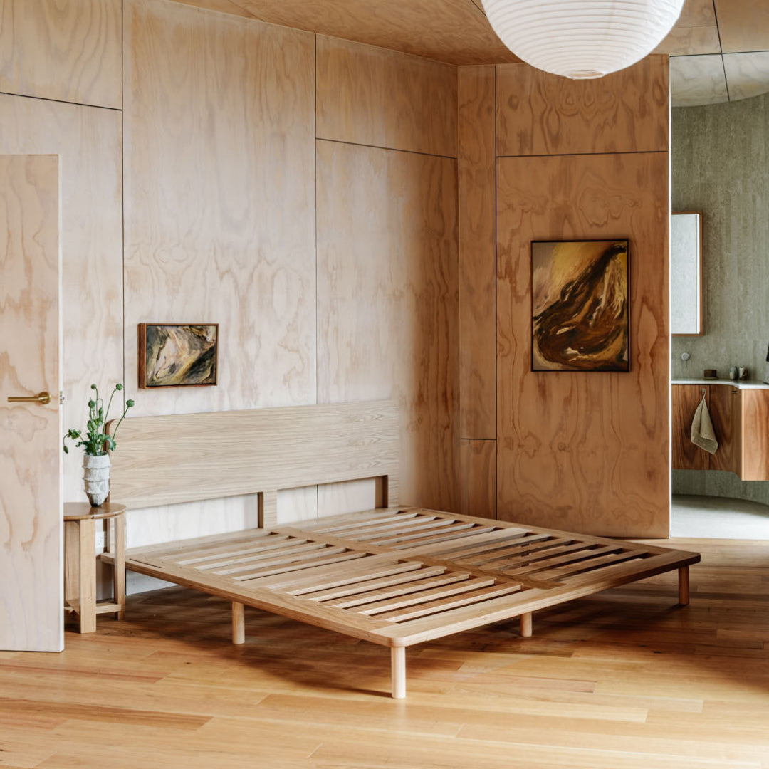 A minimalist light oak wooden bed frame with a slatted platform base and matching headboard, styled in a modern, warm-toned bedroom with plywood walls and wood flooring.